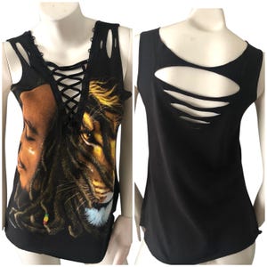 May include: Black tank top with a graphic print of a lion and a person with dreadlocks. The front features a lace-up neckline and cut-out details. The back has three horizontal cut-out slits.