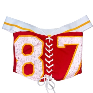 May include: Red off-the-shoulder top with the number 87 in white and yellow. The top has a white neckline with yellow stripes and a white lace-up front. The garment appears to be made of a stretchy material.