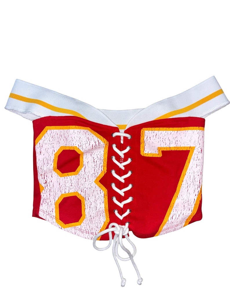May include: Red off-the-shoulder top with the number 87 in white and yellow. The top has a white neckline with yellow stripes and a white lace-up front. The garment appears to be made of a stretchy material.