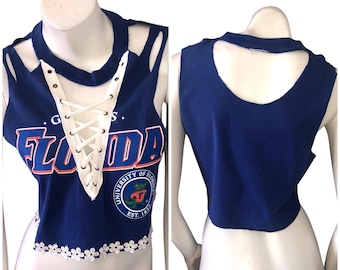 May include: A navy blue crop top with a white lace-up front and floral trim. The top features the word "FLORIDA" in orange and white, along with a university logo. The back of the top has a keyhole cutout.