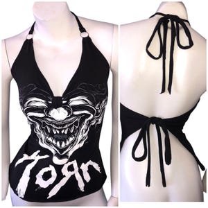 K&ouml;nnte beinhalten: Schwarzes Halstuch-Top mit einem wei&szlig;en Motiv eines gruseligen Clownsgesichts und dem Wort "Torn" auf der Vorderseite. Das Oberteil hat einen R&uuml;ckenausschnitt.