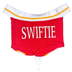 May include: Red crop top with the word "SWIFTIE" in white block letters. The top has a white elastic band with yellow stripes at the top and a drawstring at the bottom. The bottom hem has a yellow and orange pattern.