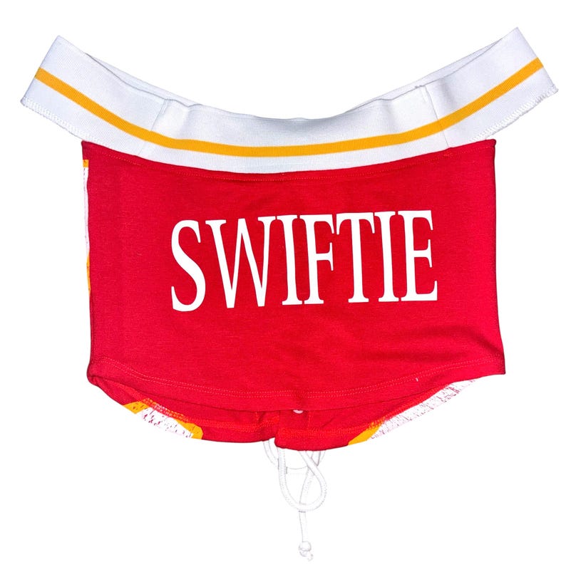May include: Red crop top with the word "SWIFTIE" in white block letters. The top has a white elastic band with yellow stripes at the top and a drawstring at the bottom. The bottom hem has a yellow and orange pattern.