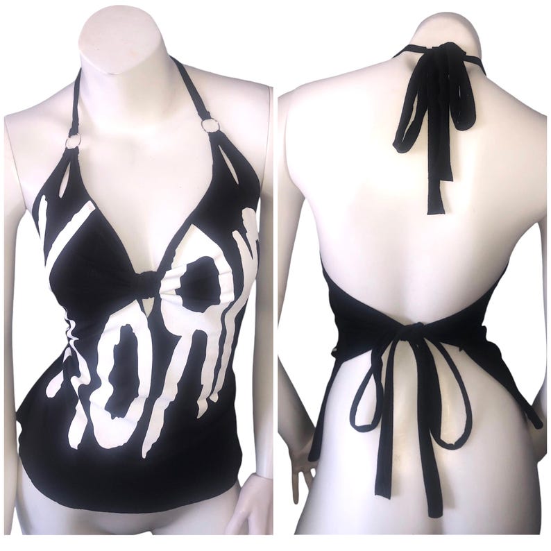 May include: Black halter top with white graphic text. The top has a plunging neckline and a tie closure at the back. The halter straps are secured with silver rings.