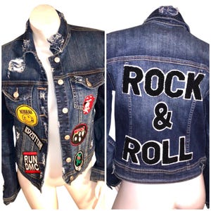 May include: A distressed blue denim jacket adorned with patches from bands like Nirvana and Led Zeppelin. The back of the jacket displays the words "ROCK & ROLL" in large, black, textured lettering.