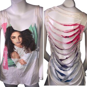 May include: White tank top featuring a portrait of a woman with dark curly hair. The front of the shirt has a colorful design with pink and green accents. The back of the shirt has a unique cut-out design with vertical slits.