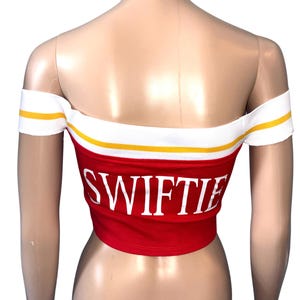 May include: Red off-the-shoulder crop top with the word "SWIFTIE" in white block letters. The top has a white band with yellow stripes along the top edge and around the shoulders. The garment is displayed on a mannequin.