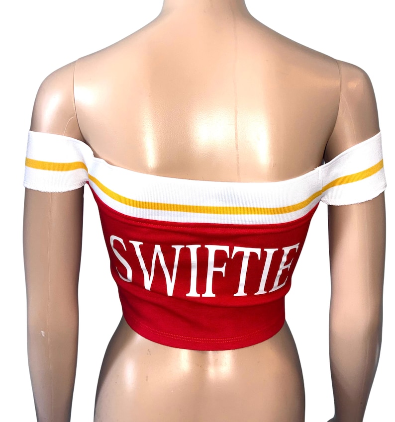 May include: Red off-the-shoulder crop top with the word "SWIFTIE" in white block letters. The top has a white band with yellow stripes along the top edge and around the shoulders. The garment is displayed on a mannequin.