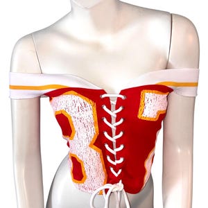 May include: Red off-the-shoulder corset top with white and yellow accents. Features the number 87 in white with yellow trim. The top has a white lace-up closure and white shoulder straps.