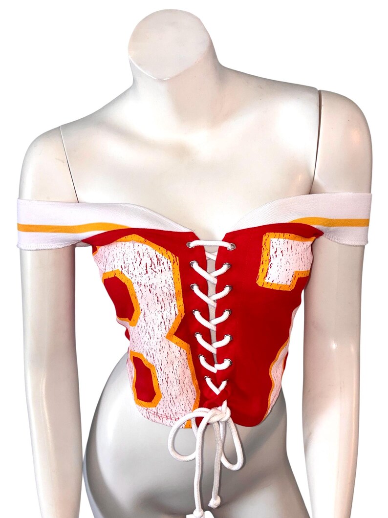May include: Red off-the-shoulder corset top with white and yellow accents. Features the number 87 in white with yellow trim. The top has a white lace-up closure and white shoulder straps.