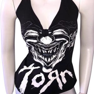 May include: Black halter top featuring a white graphic of a grinning clown face. The word "Toяn" is printed in white at the bottom. The top has a deep V-neck and is displayed on a mannequin.