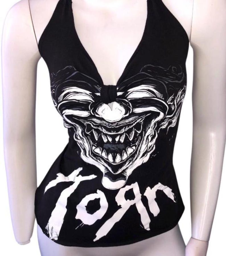 May include: Black halter top featuring a white graphic of a grinning clown face. The word "Toяn" is printed in white at the bottom. The top has a deep V-neck and is displayed on a mannequin.
