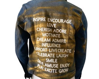 Könnte beinhalten: Eine Jeansjacke in Blau mit einem goldfarbenen Wascheffekt. Auf der Rückseite der Jacke befindet sich weißer Text mit inspirierenden Wörtern wie "Inspire", "Love" und "Smile". Klassischer Kragen und Knopfverschluss.
