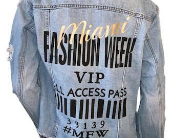 Puede incluir: Chaqueta vaquera azul claro con detalles desgastados. La parte posterior de la chaqueta presenta un gráfico dorado y negro que dice "Miami Fashion Week VIP All Access Pass" con un código de barras y el hashtag #MFW.