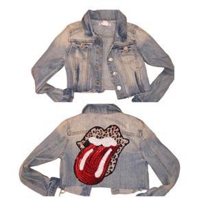 May include: A light blue denim jacket with a red, white, and black sequined tongue patch on the back. The tongue patch has a leopard print design around the edges.