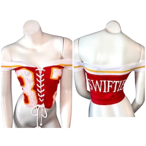May include: Red off-the-shoulder corset top with white and yellow trim. The front features the number 87 in white with yellow outline, and a white lace-up closure. The back of the top has the word "SWIFTIE" in white.