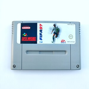 Könnte beinhalten: Eine graue Super Nintendo Entertainment System-Spielkassette für FIFA 97. Die Kassette hat ein schwarzes Etikett mit dem Spieltitel und einem Bild eines Fußballspielers.