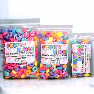 Może przedstawiać: Trzy torebki kolorowych wosków sojowych w kształcie pierścieni zbożowych. Torebki są oznaczone "Fruity Rings" i zawierają napis "Soy Wax Melts/Candle Fills" oraz "Nie jeść". Torebki mają pojemność 255 g, 170 g i 140 g.