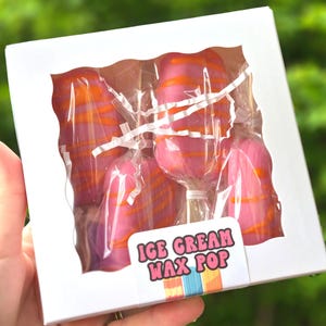 May include: A white box with a clear window displays several pink and orange swirled wax pops on sticks. The box has a sticker that reads "ICE CREAM WAX POP" in pink and blue lettering.