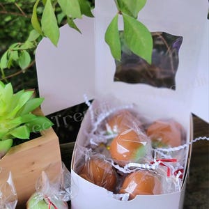 May include: A white box filled with individually wrapped caramel apples. The apples are a light green color and are covered in a layer of caramel. The box is sitting on a wooden surface.