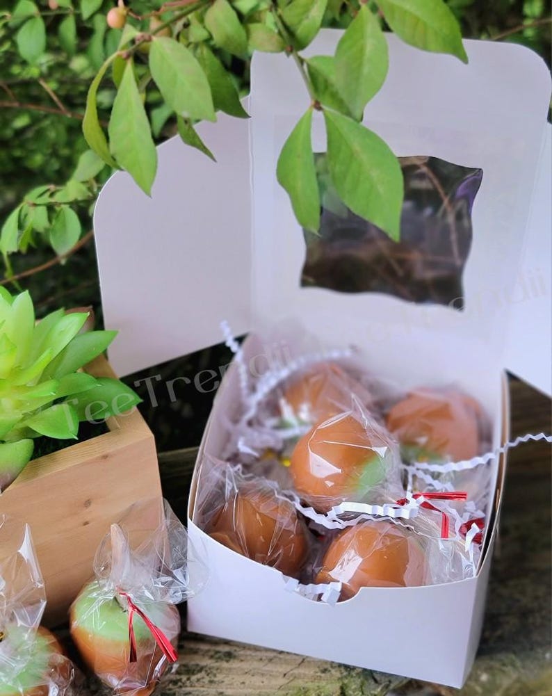 May include: A white box filled with individually wrapped caramel apples. The apples are a light green color and are covered in a layer of caramel. The box is sitting on a wooden surface.
