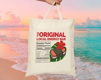 May include: A white tote bag with a red and black graphic of a Hawaiian food item called a "Spam Musubi" with the text "The Original Local Energy Bar" and "Kaukau Facts" with nutritional information. The bag is being held by a hand against a background of a beach and ocean.
