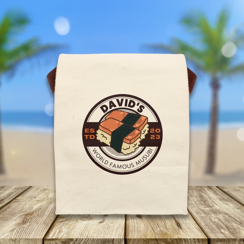 May include: A white canvas tote bag with brown straps and a brown circle logo. The logo says "David's World Famous Musubi" and features a cartoon image of two musubi, a Hawaiian dish of rice and spam wrapped in seaweed. The logo also includes the text "Est. 2023".