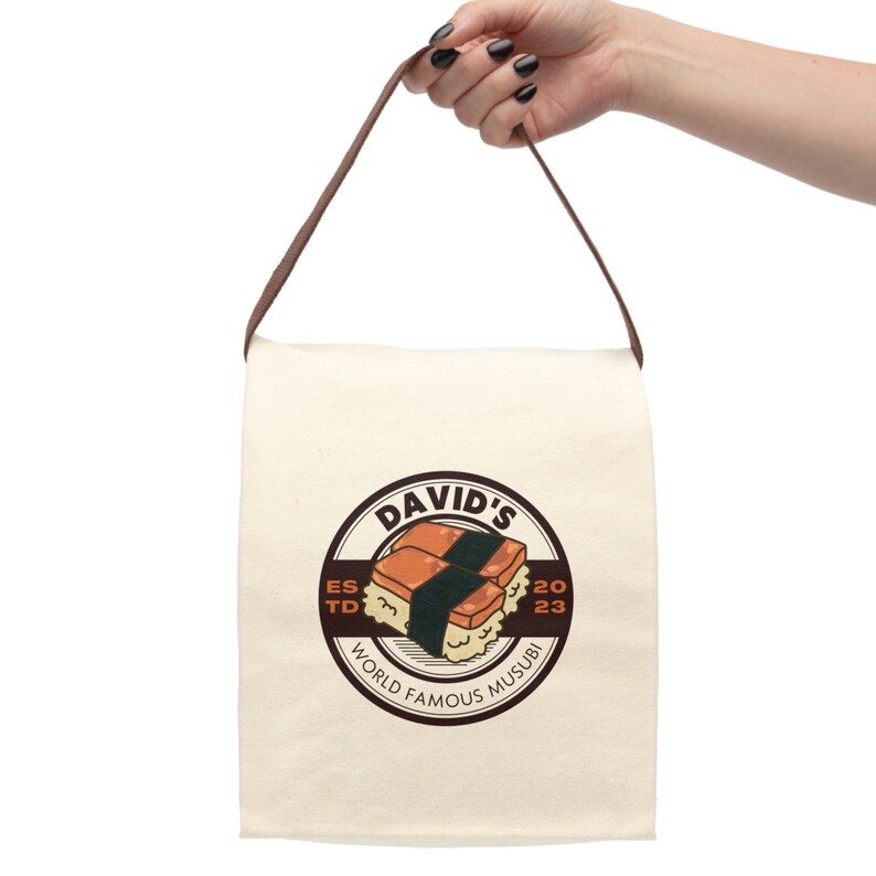 May include: A white canvas tote bag with brown straps. The bag has a brown circle logo with the text "DAVID'S WORLD FAMOUS MUSUBI" and a cartoon image of two musubi.