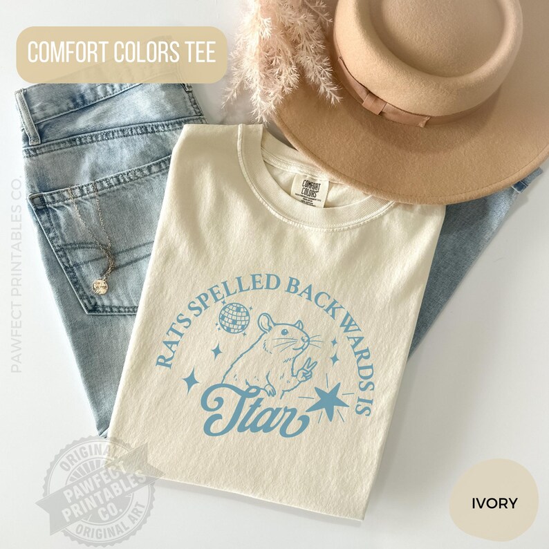 May include: Ivory-colored Comfort Colors tee with the text "Rats Spelled Backwards Is Star" in light blue. The graphic includes a rat, a disco ball, and stars. The shirt is paired with blue jeans and a tan hat.