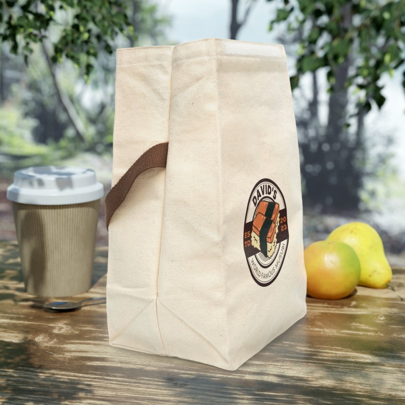 May include: A beige canvas lunch bag with a brown leather handle and a logo that says "David's Est'd 2023 World Famous Musubi".