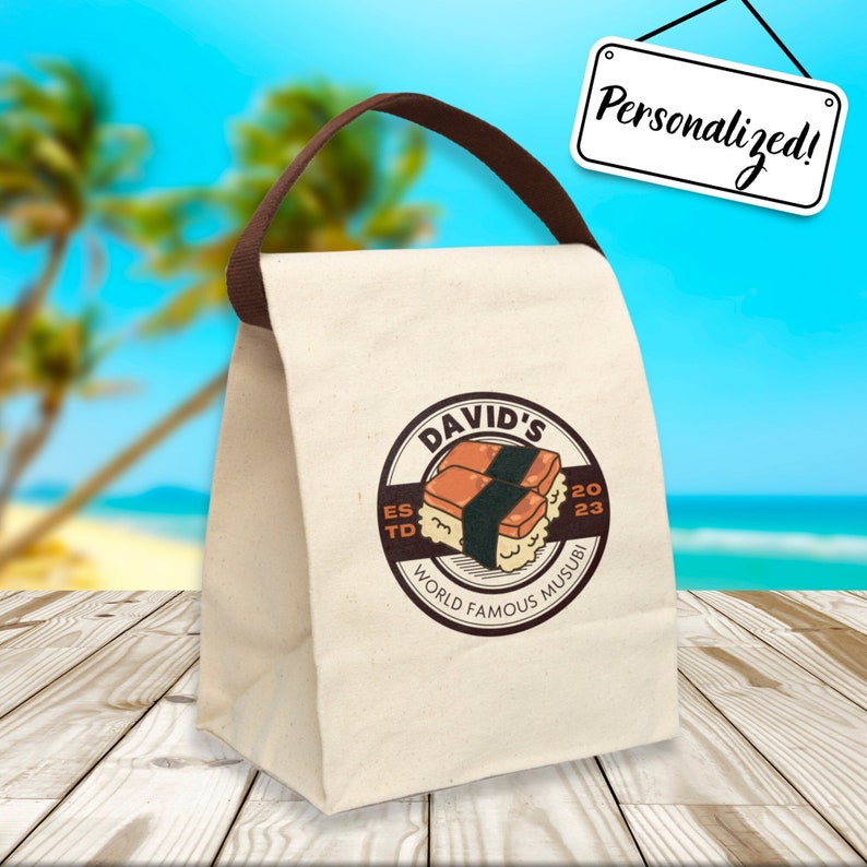 May include: A white canvas lunch bag with a brown handle. The bag has a logo that says "David's World Famous Musubi Est. 2023" with a cartoon image of two musubi.