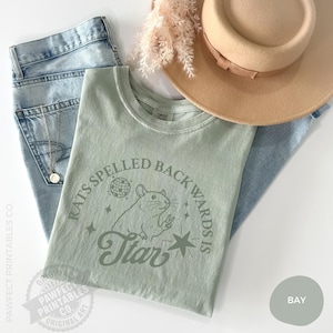 May include: A light green t-shirt with the text "Rats Spelled Backwards Is Star" and a rat illustration. The shirt is paired with light blue jeans, a gold necklace, and a tan hat. The shirt is a Comfort Colors brand.