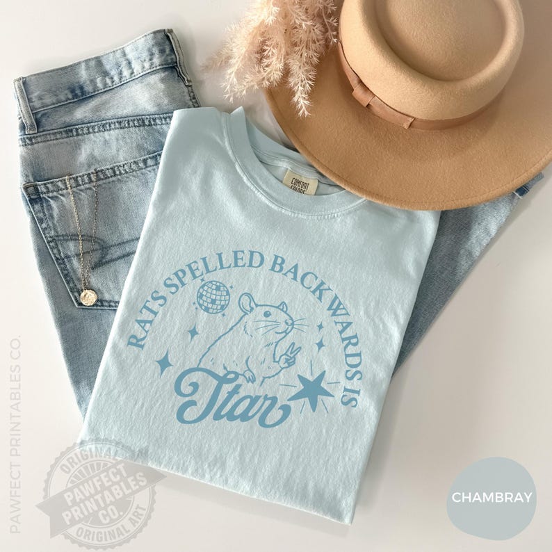 May include: A light blue t-shirt with the text "Rats Spelled Backwards is Star" and a rat graphic. The shirt is paired with light wash jeans and a tan hat. A gold necklace is visible.