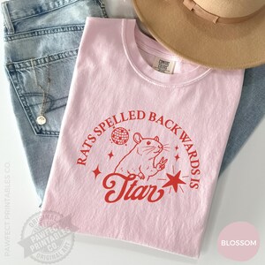 May include: A light pink t-shirt with the text "RATS SPELLED BACKWARDS IS STAR" in red. The graphic includes a rat making a peace sign, a disco ball, and stars. The shirt is folded on top of blue jeans and a tan hat.