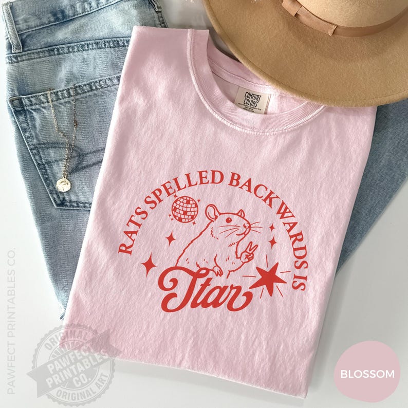 May include: A light pink t-shirt with the text "RATS SPELLED BACKWARDS IS STAR" in red. The graphic includes a rat making a peace sign, a disco ball, and stars. The shirt is folded on top of blue jeans and a tan hat.