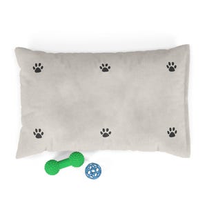 May include: A rectangular, light beige pet bed with black paw prints scattered across the surface. A green dumbbell-shaped toy and a blue ball toy are placed near the bed, suggesting a pet-friendly theme.