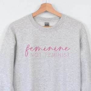 K&ouml;nnte beinhalten: Grauer Pullover mit dem Text "feminine not feminist" in rosa Schrift.