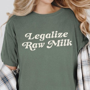 May include: A moss green t-shirt with the text "Legalize Raw Milk" in a retro font. The shirt is short-sleeved and has a relaxed fit. A plaid shirt is draped over the shoulders.