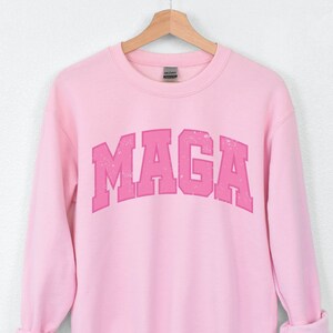 May include: Pink sweatshirt with a pink "MAGA" graphic printed on the front.