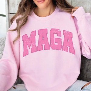 May include: A light pink sweatshirt with the text "MAGA" printed in a distressed, varsity-style font.