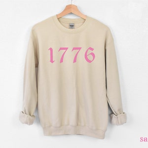 May include: A beige sweatshirt with a pink 1776 printed on the front in a gothic font.