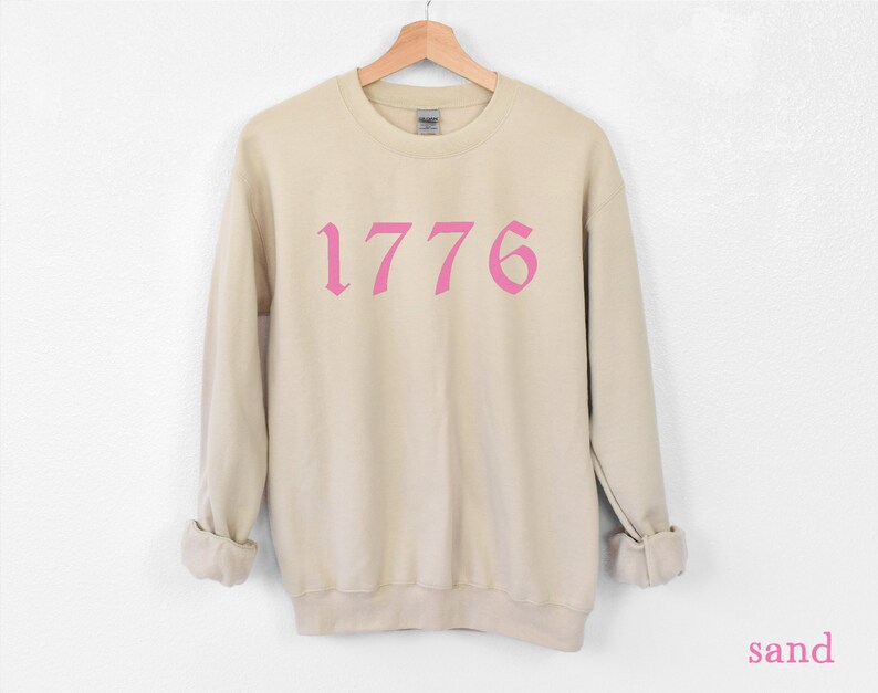 May include: A beige sweatshirt with a pink 1776 printed on the front in a gothic font.