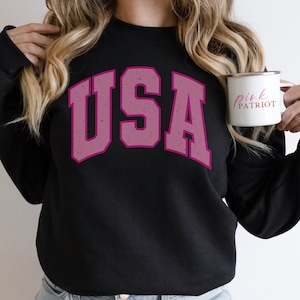 以下が含まれることがあります： 色あせたピンク色の文字で「USA」と書かれた黒いスウェットシャツ。ピンク色の「Pink Patriot」という文字が入った白いマグカップが手に持たれています。