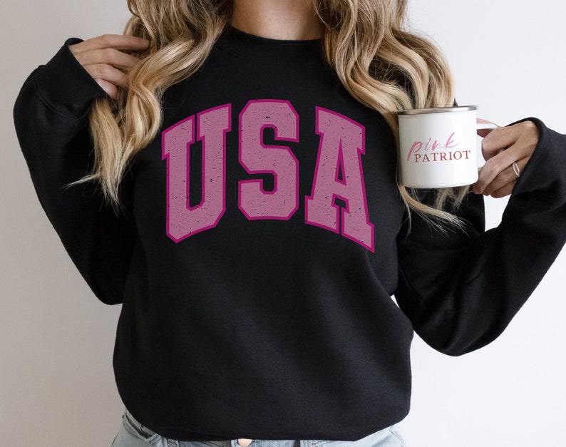 以下が含まれることがあります： 色あせたピンク色の文字で「USA」と書かれた黒いスウェットシャツ。ピンク色の「Pink Patriot」という文字が入った白いマグカップが手に持たれています。
