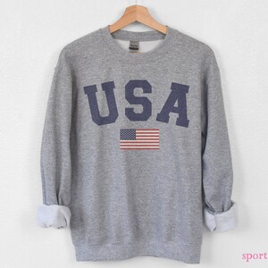 Op de afbeelding: Een grijs sweatshirt met het woord "USA" in blauw gedrukt op de voorkant. Een kleine Amerikaanse vlag is gedrukt onder het woord. Het sweatshirt is een ronde hals stijl.