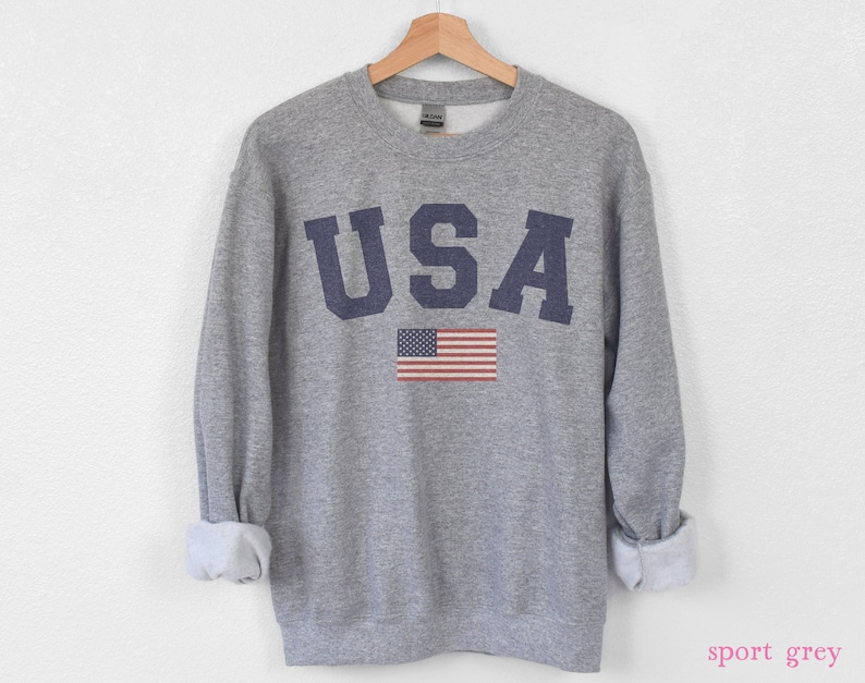 Op de afbeelding: Een grijs sweatshirt met het woord "USA" in blauw gedrukt op de voorkant. Een kleine Amerikaanse vlag is gedrukt onder het woord. Het sweatshirt is een ronde hals stijl.