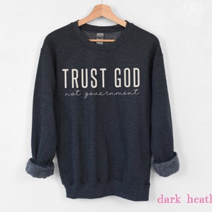 以下が含まれることがあります： 濃いグレーのヘザーのトレーナーに、白い文字で「TRUST GOD not government」と書いてあります。