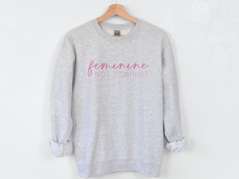 K&ouml;nnte beinhalten: Grauer Pullover mit dem Text "feminine not feminist" in rosa Schrift.