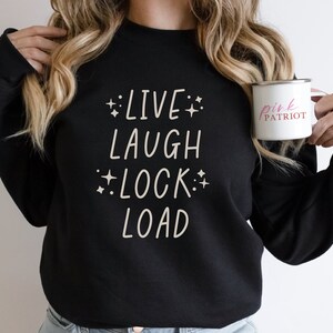 May include: Black sweatshirt with the text "LIVE LAUGH LOCK LOAD" in white with small stars around the words. A person is holding a pink enamel mug with the text "pink PATRIOT" in white.