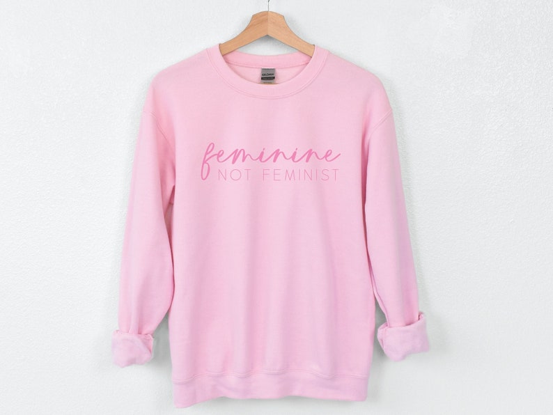 K&ouml;nnte beinhalten: Rosafarbener Pullover mit dem Text "feminine not feminist" in rosa Schrift.